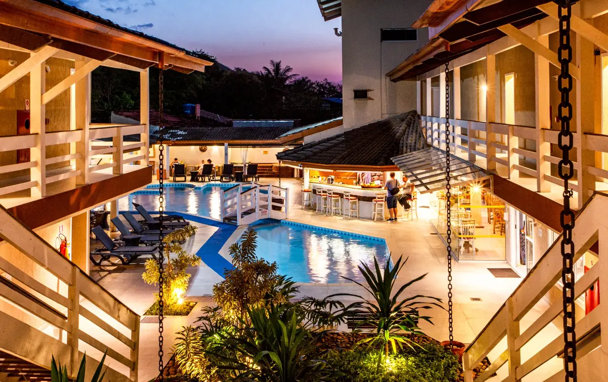 Área de lazer do hotel ao entardecer, com piscinas iluminadas, espreguiçadeiras e bar cercados pelas varandas dos apartamentos