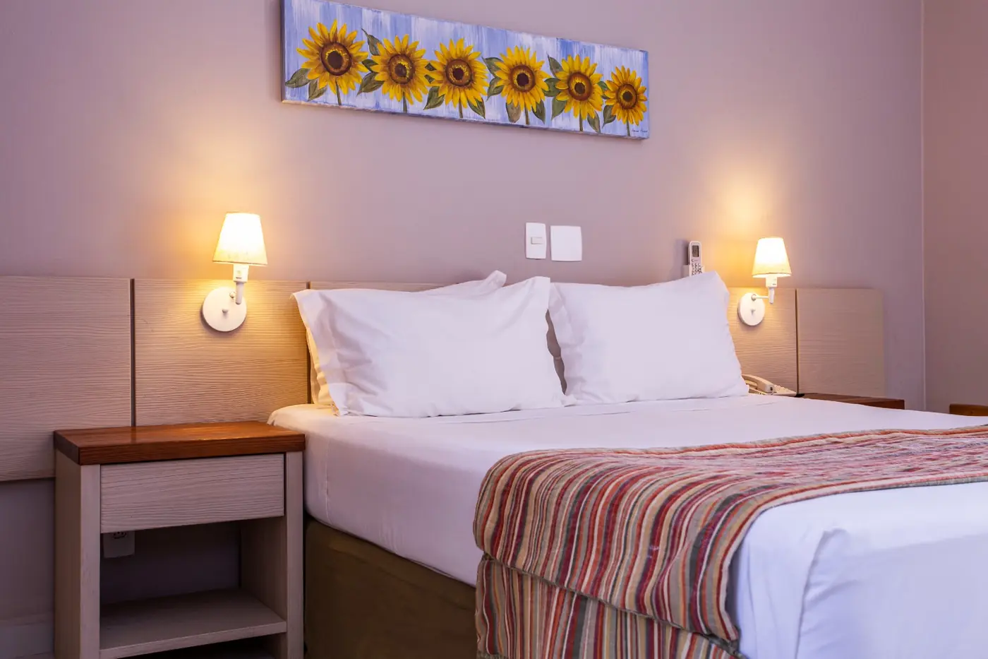 Apartamento com cama de casal arrumada com lençóis brancos, manta listrada, arandelas acesas e quadro de girassóis